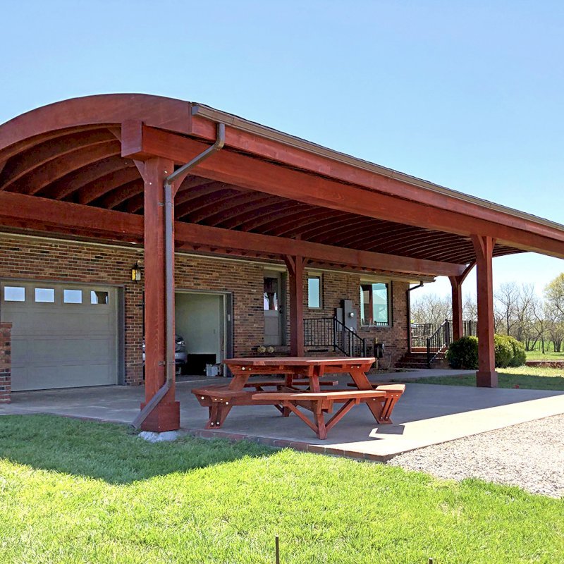 Custom Arched Thick Timber Pavilion (Options: 55' L x 22' Arc W, California Redwood, No Electrical Wiring Trim, 6 Post Anchor Kit for Gale-Wind, 2 Ceiling Fan Bases, No Privacy Panel, No Curtain Rods, 10ft H, Cherry Stain Premium Sealant) Support timbers covered by custom request. Photo also shows a Round Picnic Table. Photo Courtesy of J. Hartmann of Pittsburg, KS. Custom Arched Thick Timber Pavilion (Options: 55' L x 22' Arc W, California Redwood, No Electrical Wiring Trim, 6 Post Anchor Kit for Gale-Wind, 2 Ceiling Fan Bases, No Privacy Panel, No Curtain Rods, 10ft H, Cherry Stain Premium Sealant) Support timbers covered by custom request. Photo also shows a Round Picnic Table. Photo Courtesy of J. Hartmann of Pittsburg, KS.