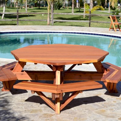 Octagonal Picnic Table (Options: 6' Diameter Tabletop, Attached Benches, Mature Redwood, Standard Tabletop, Rounded Corners, No Umbrella Hole, Transparent Premium Sealant). Round Folding Table with Chairs in Background. 
