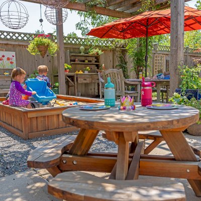 Round Wooden Picnic Table for Toddlers (Mosaic Eco-Wood, No Umbrella Hole, Standard Tabletop, Transparent Premium Sealant). Photo Also Shows a La Playa Sandbox and an Eli's Potting Bench. Photo Courtesy of M. Cooke of Westchester, IL.