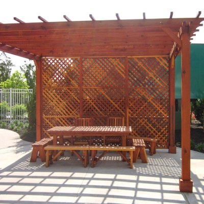 Custom Traditional Wooden Garden Pergola (Options: 15' x 15', California Redwood, No Electrical Wiring Trim, Open Roof with Slats at 18", Rafters at 18", 4-Post Anchor Kit for Stone, No Ceiling Fan Base, 1 Privacy Panel [1 full side enclosed by custom request], No Curtain Rods, 9' Post Height, Transparent Premium Sealant). Under the Pergola, a Forever Picnic Table with Unattached Benches and 2 Ruth Chairs. Photo Courtesy of Susannah Shaw of New York, NY.