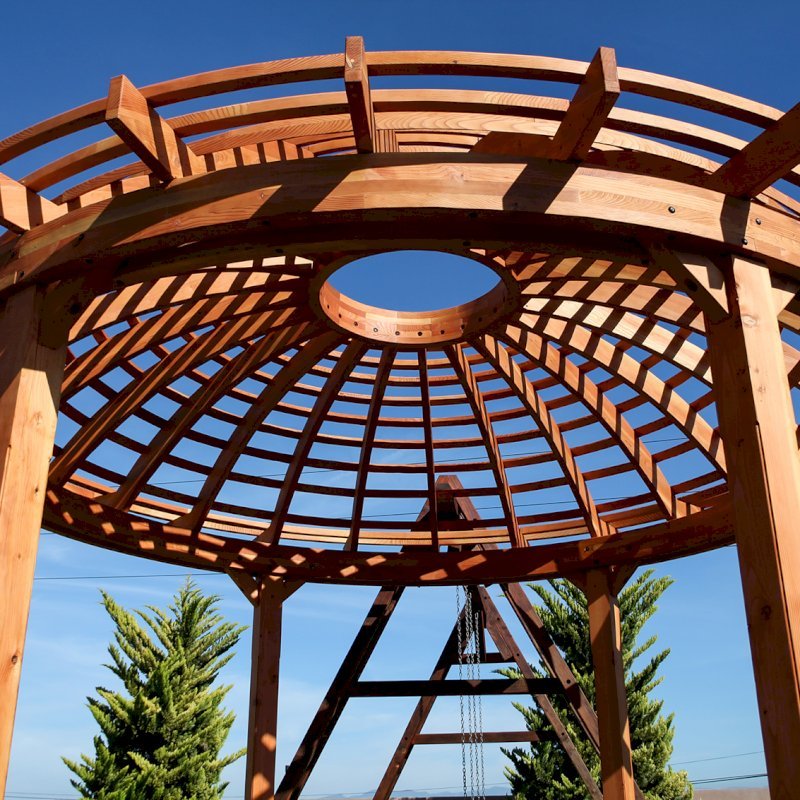 The Wooden Dome Pergola (Options: 16' Diameter, with Deck in Coffee Stain, Douglas-fir, Open Roof with Slats at 6", 9' Posts, No Electrical Wiring Trim, 6-Post Anchor Kit for Concrete, Transparent Premium Sealant). Photo Also shows a Round Picnic Table and a Rory's Swing Set in Background. The Wooden Dome Pergola (Options: 16' Diameter, with Deck in Coffee Stain, Douglas-fir, Open Roof with Slats at 6", 9' Posts, No Electrical Wiring Trim, 6-Post Anchor Kit for Concrete, Transparent Premium Sealant). Photo Also shows a Round Picnic Table and a Rory's Swing Set in Background.