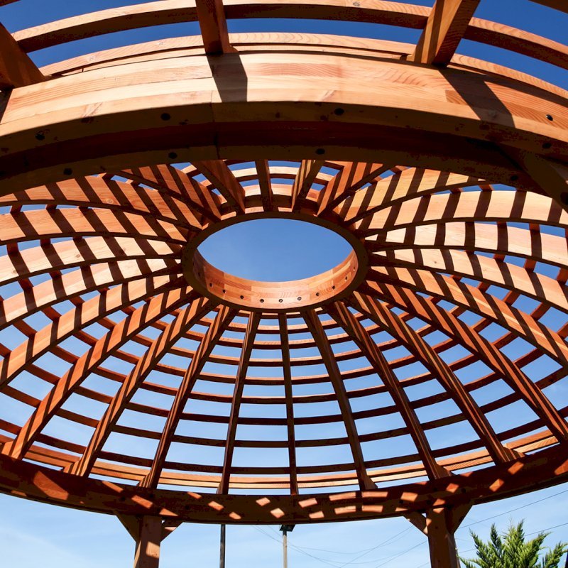 The Wooden Dome Pergola (Options: 16' Diameter, with Deck in Coffee Stain, Douglas-fir, Open Roof with Slats at 6", 9' Posts, No Electrical Wiring Trim, 6-Post Anchor Kit for Concrete, Transparent Premium Sealant). Photo Also shows a Round Picnic Table and a Rory's Swing Set in Background. The Wooden Dome Pergola (Options: 16' Diameter, with Deck in Coffee Stain, Douglas-fir, Open Roof with Slats at 6", 9' Posts, No Electrical Wiring Trim, 6-Post Anchor Kit for Concrete, Transparent Premium Sealant). Photo Also shows a Round Picnic Table and a Rory's Swing Set in Background.