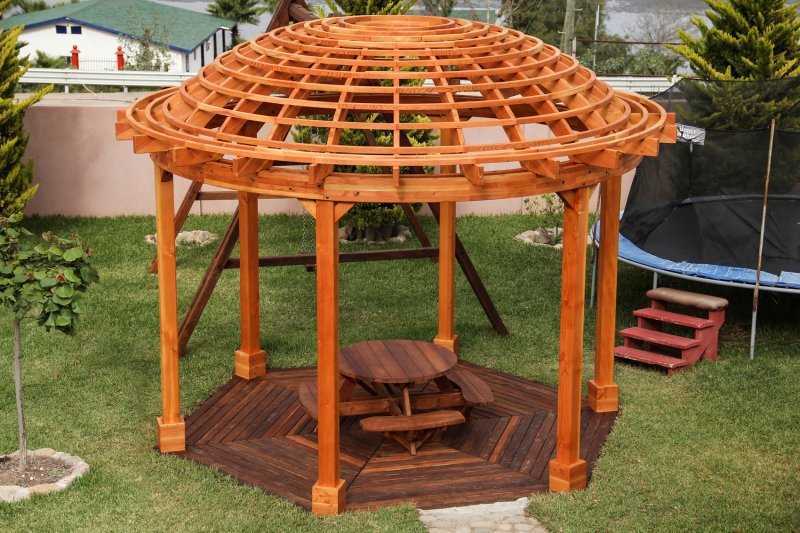 The Wooden Dome Pergola (Options: 16' Diameter, with Deck in Coffee Stain, Douglas-fir, Open Roof with Slats at 6", 9' Posts, No Electrical Wiring Trim, 6-Post Anchor Kit for Concrete, Transparent Premium Sealant). Photo Also shows a Round Picnic Table and a Rory's Swing Set in Background. The Wooden Dome Pergola (Options: 16' Diameter, with Deck in Coffee Stain, Douglas-fir, Open Roof with Slats at 6", 9' Posts, No Electrical Wiring Trim, 6-Post Anchor Kit for Concrete, Transparent Premium Sealant). Photo Also shows a Round Picnic Table and a Rory's Swing Set in Background.