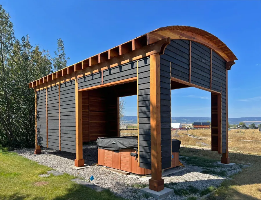 Custom  Arched Thick Timber Pavilion (Options: 28' L x 18' Arc W, California Redwood, Post Anchor Kit for High-Wind, with Custom Privacy Walls, 10' & 12' H Posts, No Curtain Rods, Walls Siding with Black Stain and the Rest of the Structure with Transparent Premium Sealant). Photo Courtesy of D. Harris of Tetonia, ID.