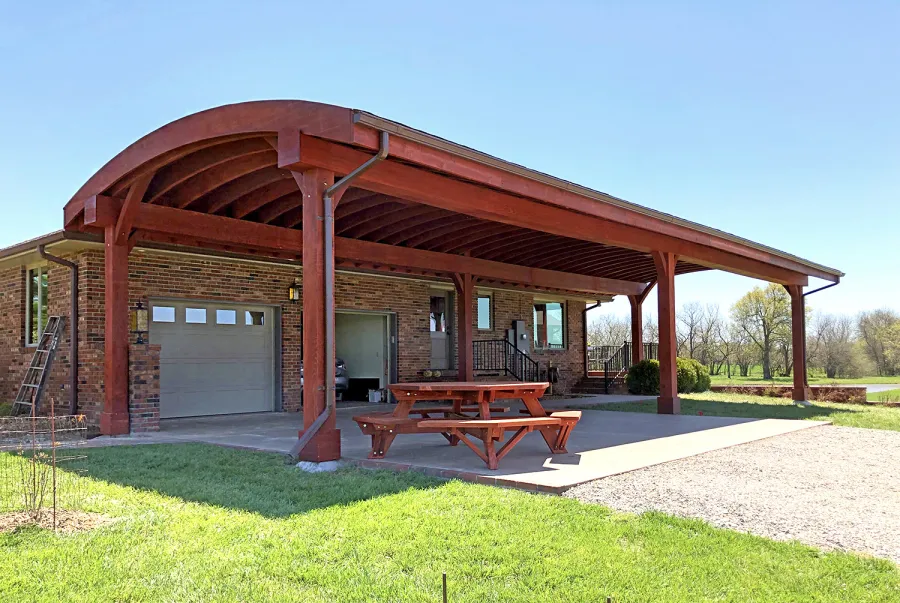 Custom Arched Thick Timber Pavilion (Options: 55' L x 22' Arc W, California Redwood, No Electrical Wiring Trim, 6-Post Anchor Kit for High-Wind, 2 Ceiling Fan Bases, No Privacy Panel, No Curtain Rods, 10ft H, Cherry Stain Premium Sealant) Support timbers covered by custom request. Photo also shows a Round Picnic Table. Photo Courtesy of J. Hartmann of Pittsburg, KS.