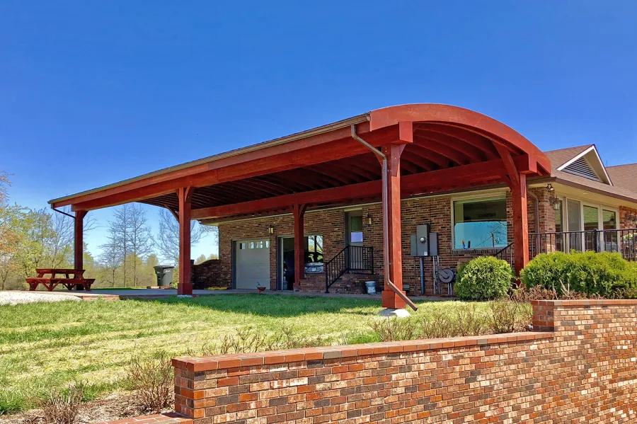 Custom Arched Thick Timber Pavilion (Options: 55' L x 22' Arc W, California Redwood, No Electrical Wiring Trim, 6-Post Anchor Kit for High-Wind, 2 Ceiling Fan Bases, No Privacy Panel, No Curtain Rods, 10ft H, Cherry Stain Premium Sealant) Support timbers covered by custom request. Photo also shows a Round Picnic Table. Photo Courtesy of J. Hartmann of Pittsburg, KS.