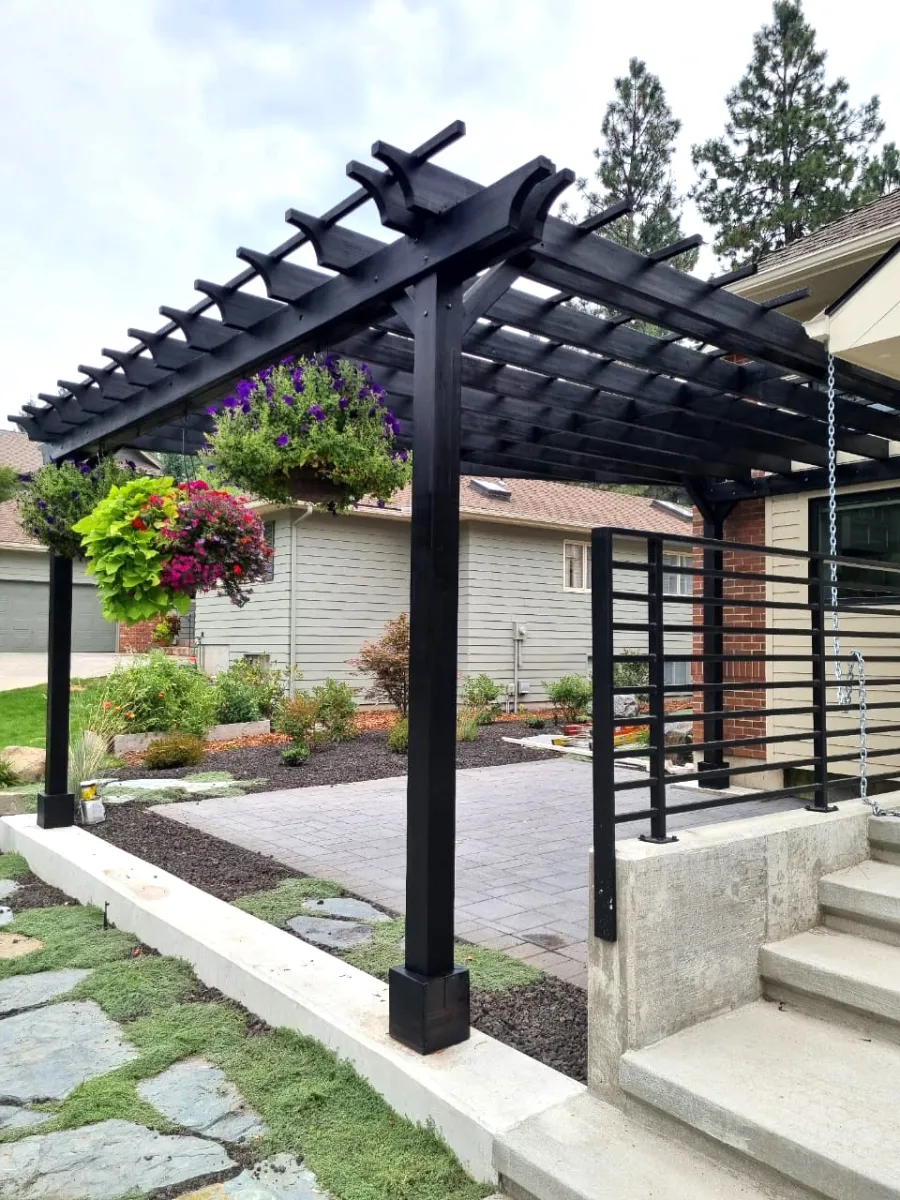 Custom Attached Garden Perglola (Options: Trapezoid Roof Shape [19' Front, 12' Back, 20' Deep], Mosaic Douglas-Fir, Open Roof with Slats at 18", Rafters at 18", with 3 Posts by Custom Request, No Rain Guard, 3-Post Anchor Kit for Concrete, No Privacy Panel, No Ceiling Fan Base, Black Stain Premium Sealant). Photo Taken 1 Year After Installation [After Resealed]. Photo Courtesy of T. Perko of Spokane, WA.