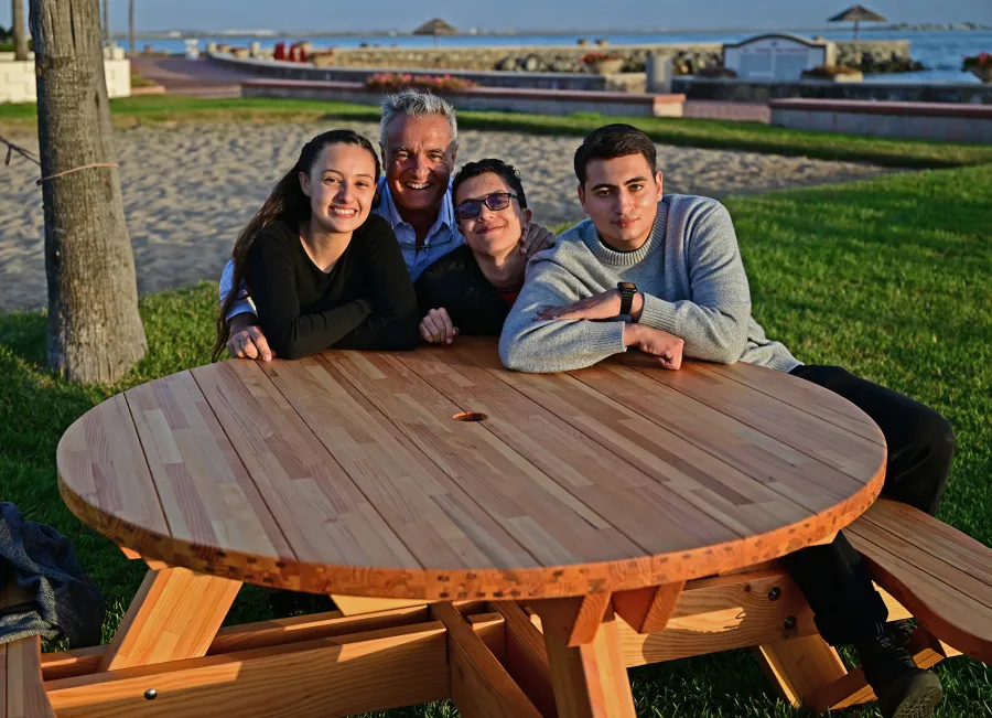 Round Wooden Picnic Table (Attached Benches) (Options: 5' Diameter, Attached Benches, Mosaic Douglas-Fir, Standard Tabletop, No Lazy Susan, 2" Umbrella Hole, Transparent Premium Sealant). Photo courtesy of El Estero Beach Resort in Ensenada, Mexico.