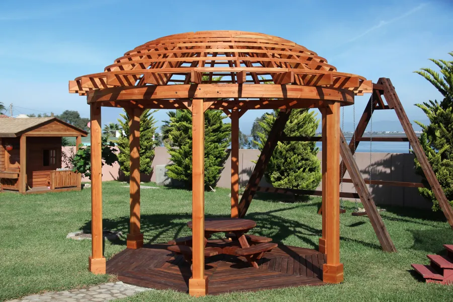 Wooden Dome Pergola (Options: 16' Diameter, with Deck in Coffee Stain, Douglas-fir, Open Roof with Slats at 6", 9' Posts, No Electrical Wiring Trim, 6-Post Anchor Kit for Concrete, Transparent Premium Sealant). Photo Also shows a Round Picnic Table and a Rory's Swing Set in Background.
