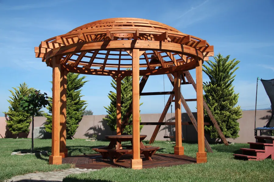 Wooden Dome Pergola (Options: 16' Diameter, with Deck in Coffee Stain, Douglas-fir, Open Roof with Slats at 6", 9' Posts, No Electrical Wiring Trim, 6-Post Anchor Kit for Concrete, Transparent Premium Sealant). Photo Also shows a Round Picnic Table and a Rory's Swing Set in Background.