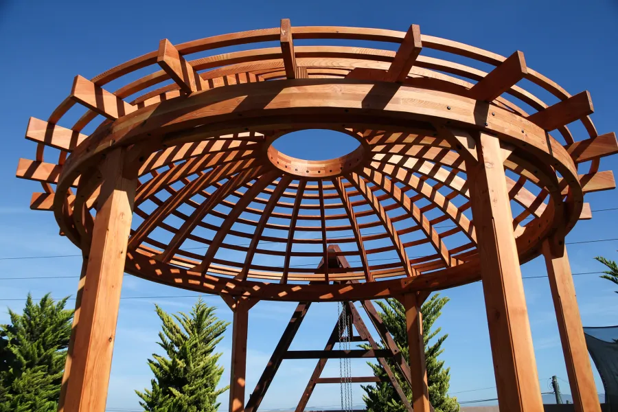Wooden Dome Pergola (Options: 16' Diameter, with Deck in Coffee Stain, Douglas-fir, Open Roof with Slats at 6", 9' Posts, No Electrical Wiring Trim, 6-Post Anchor Kit for Concrete, Transparent Premium Sealant). Photo Also shows a Round Picnic Table and a Rory's Swing Set in Background.