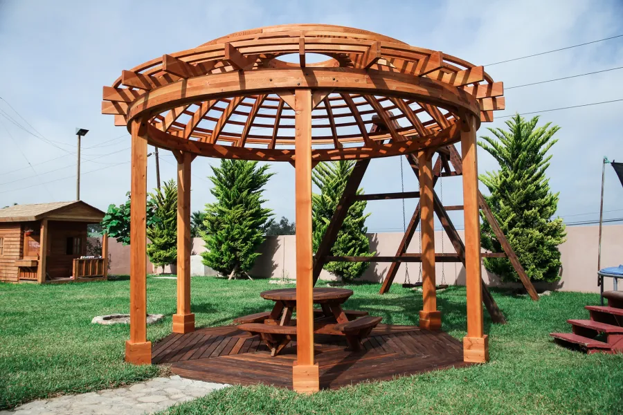 Wooden Dome Pergola (Options: 16' Diameter, with Deck in Coffee Stain, Douglas-fir, Open Roof with Slats at 6", 9' Posts, No Electrical Wiring Trim, 6-Post Anchor Kit for Concrete, Transparent Premium Sealant). Photo Also shows a Round Picnic Table and a Rory's Swing Set in Background.