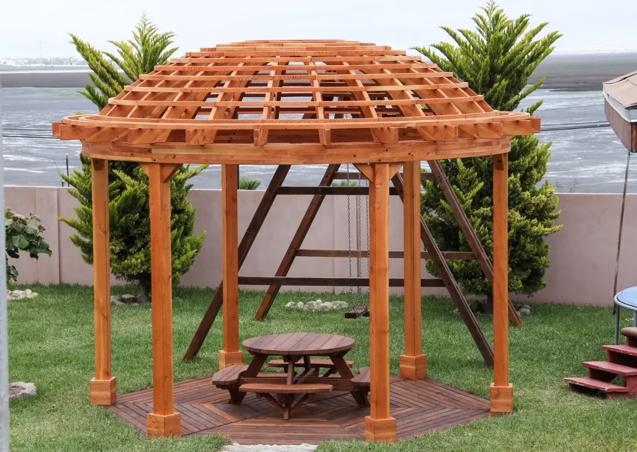 Wooden Dome Pergola (Options: 16' Diameter, with Deck in Coffee Stain, Douglas-fir, Open Roof with Slats at 6", 9' Posts, No Electrical Wiring Trim, 6-Post Anchor Kit for Concrete, Transparent Premium Sealant). Photo Also shows a Round Picnic Table and a Rory's Swing Set in Background.
