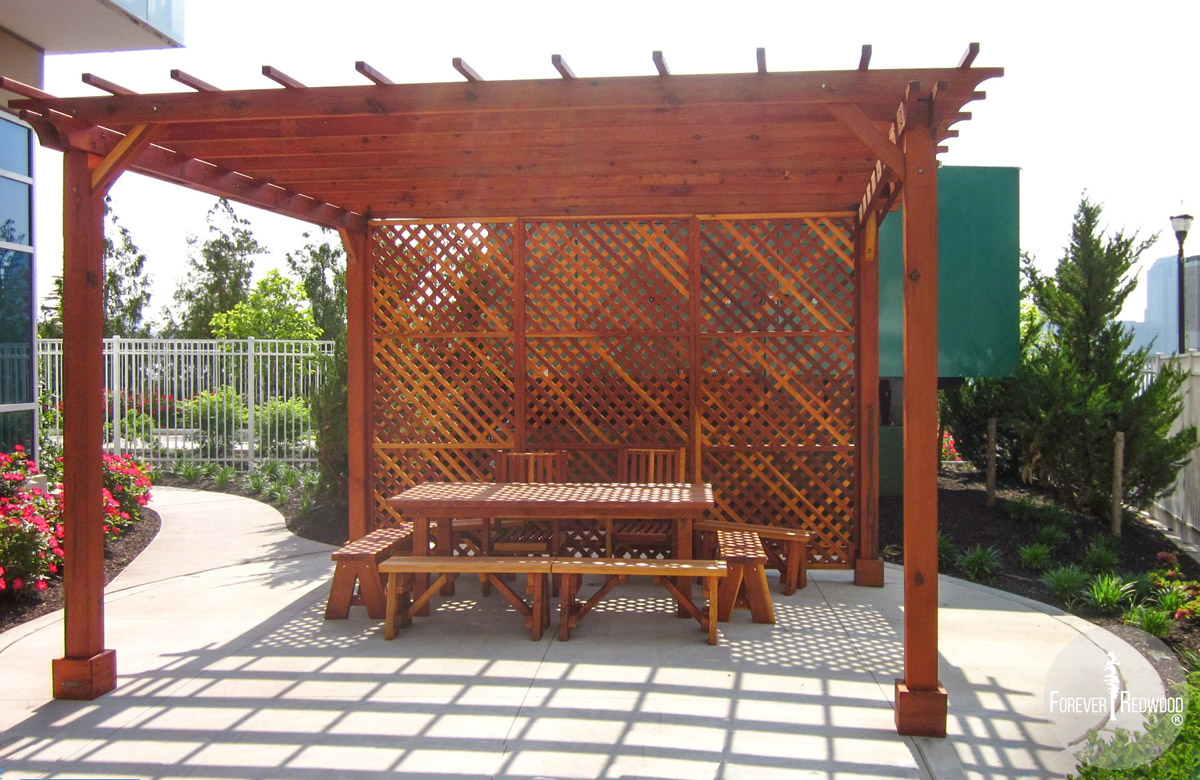 Custom Traditional Wooden Garden Pergola (Options: 15' x 15', California Redwood, No Electrical Wiring Trim, Open Roof with Slats at 18", Rafters at 18", 4-Post Anchor Kit for Stone, No Ceiling Fan Base, 1 Privacy Panel [1 full side enclosed by custom request], No Curtain Rods, 9' Post Height, Transparent Premium Sealant). Under the Pergola, a Forever Picnic Table with Unattached Benches and 2 Ruth Chairs. Photo Courtesy of Susannah Shaw of New York, NY.